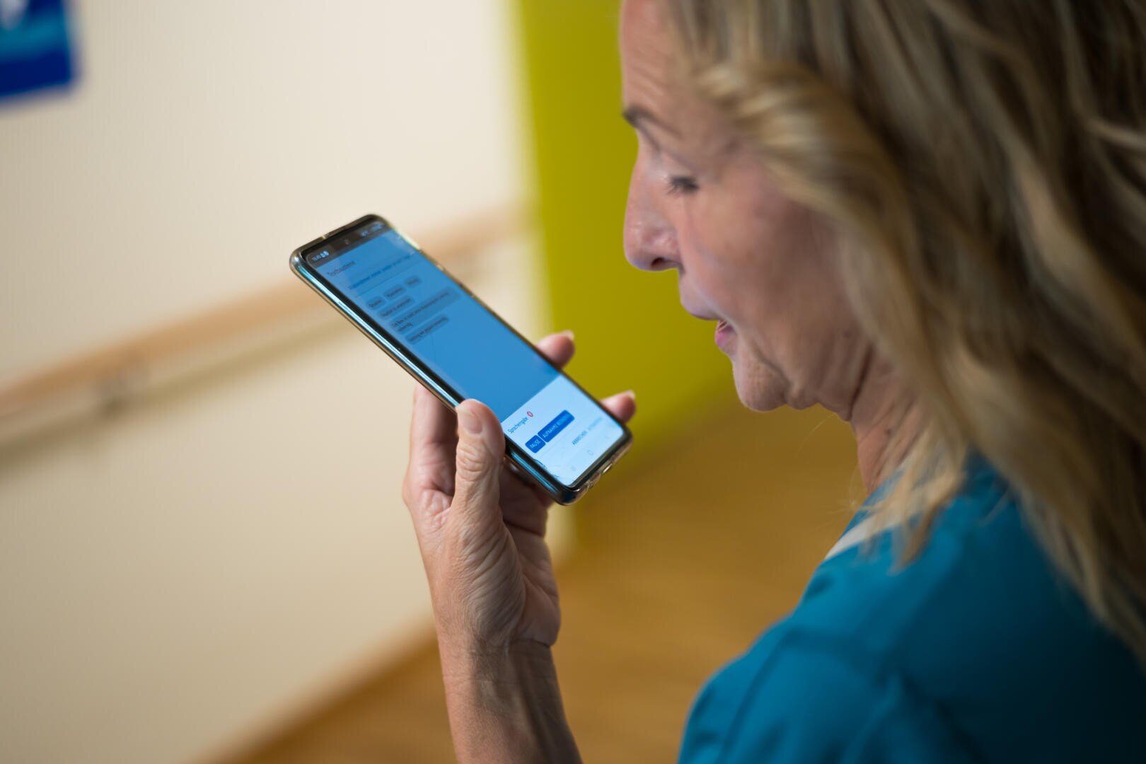 Eine Frau mit welligem blondem Haar hält ein Smartphone in der Hand und spricht hinein, wobei sie eine Sprach-zu-Text- oder Sprachassistenzfunktion nutzt. Der Hintergrund ist unscharf und zeigt eine grüne Wand und einen Holzboden.