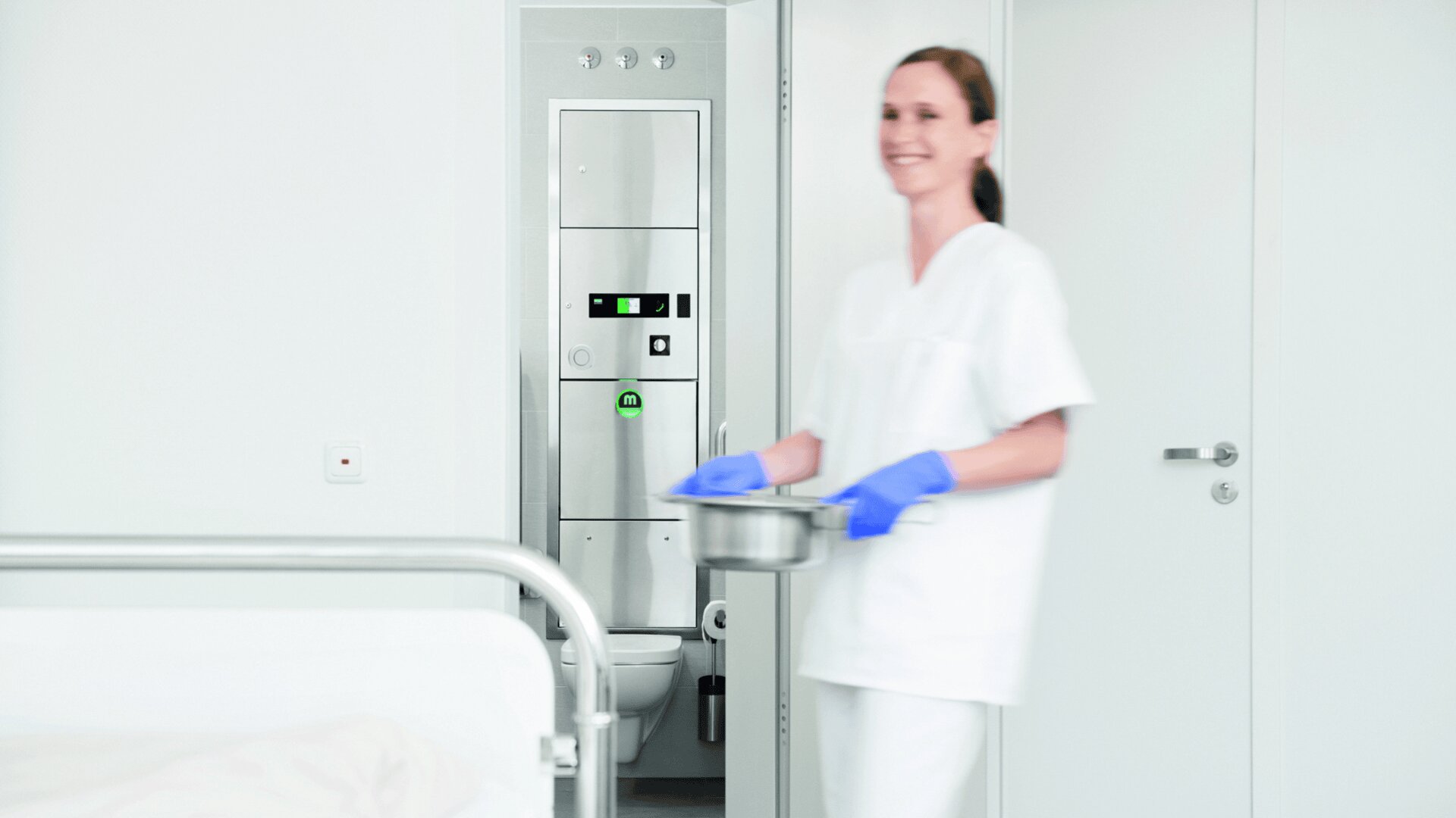 Eine Krankenschwester in weißem Kittel und blauen Handschuhen hält einen Metallbehälter und lächelt, während sie in einem sauberen Krankenhauszimmer steht, in dem ein Bett und medizinische Geräte zu sehen sind.