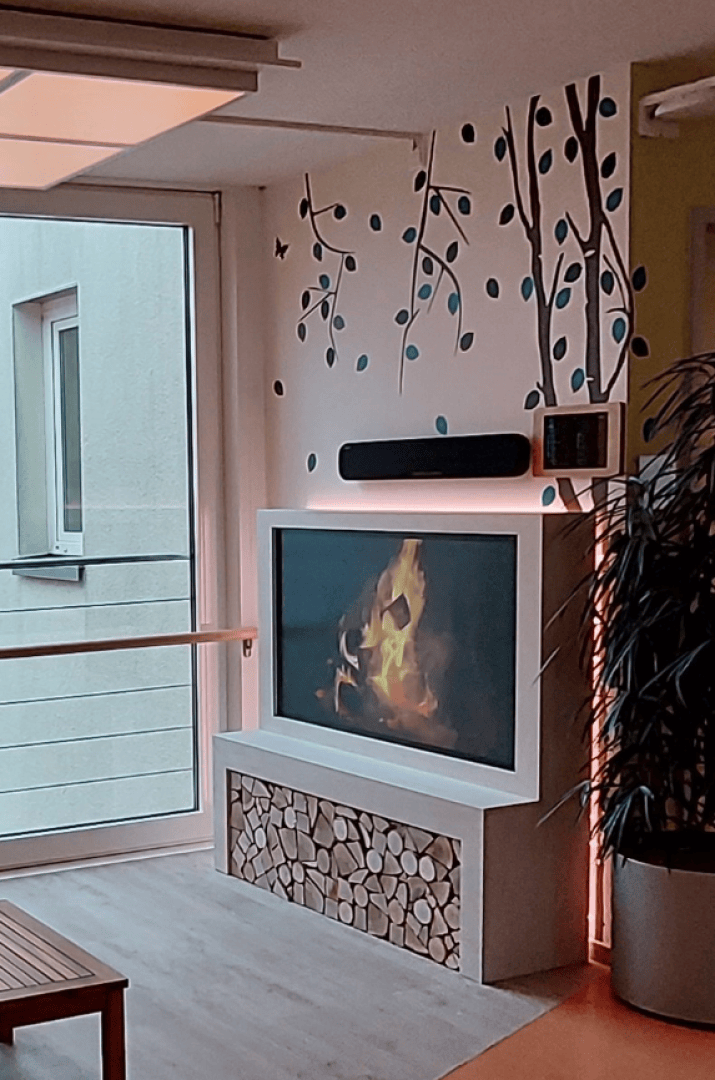 Une cheminée murale dotée d'un écran numérique reproduisant des flammes trône au-dessus d'un socle décoratif en forme de bûches de bois, posée contre un mur orné d'une fresque représentant un arbre stylisé.