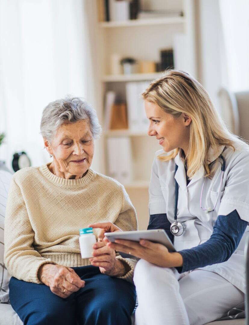 Un professionnel de santé souriant, muni d'un stéthoscope, montre une tablette à une femme âgée qui tient un flacon de médicaments. Ils sont assis ensemble dans un salon lumineux et discutent amicalement.
