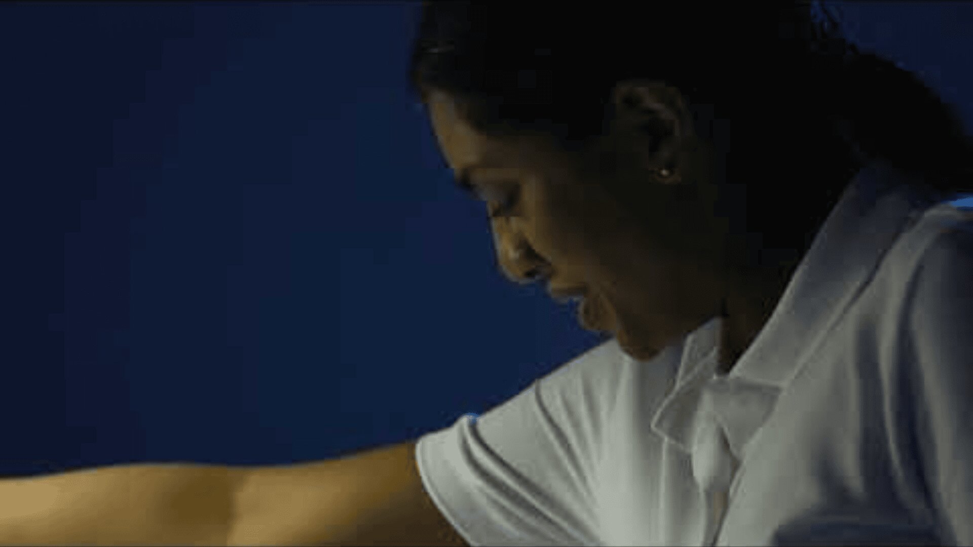 Une femme vêtue d'une chemise blanche regarde vers le bas et sourit légèrement, avec un arrière-plan bleu foncé derrière elle.