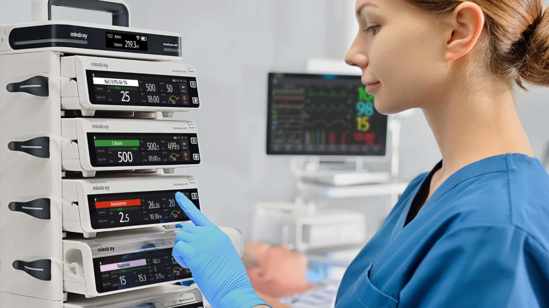 Une infirmière en blouse bleue et gants manipule une pile de pompes à perfusion médicales, avec un moniteur patient affichant les signes vitaux et un mannequin patient flou à l'arrière-plan.
