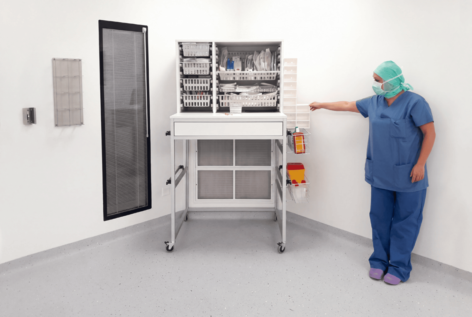 Un professionnel de santé, vêtu d'une tenue chirurgicale et portant un masque, se tient dans une pièce blanche et propre et désigne une unité d'élimination des déchets fixée au mur, à côté d'une grande armoire ouverte contenant des consommables médicaux.