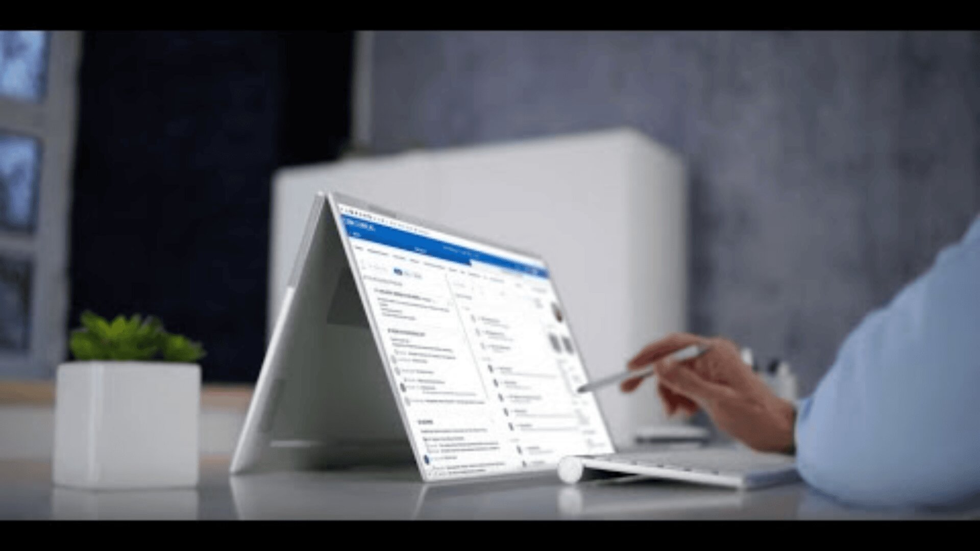 Eine Person verwendet einen Stift, um mit einem Tablet zu interagieren, das im Zeltmodus auf einem Schreibtisch installiert ist, mit einer drahtlosen Tastatur und einer kleinen Topfpflanze in der Nähe. Auf dem Bildschirm wird eine organisierte Anwendungsoberfläche angezeigt.
