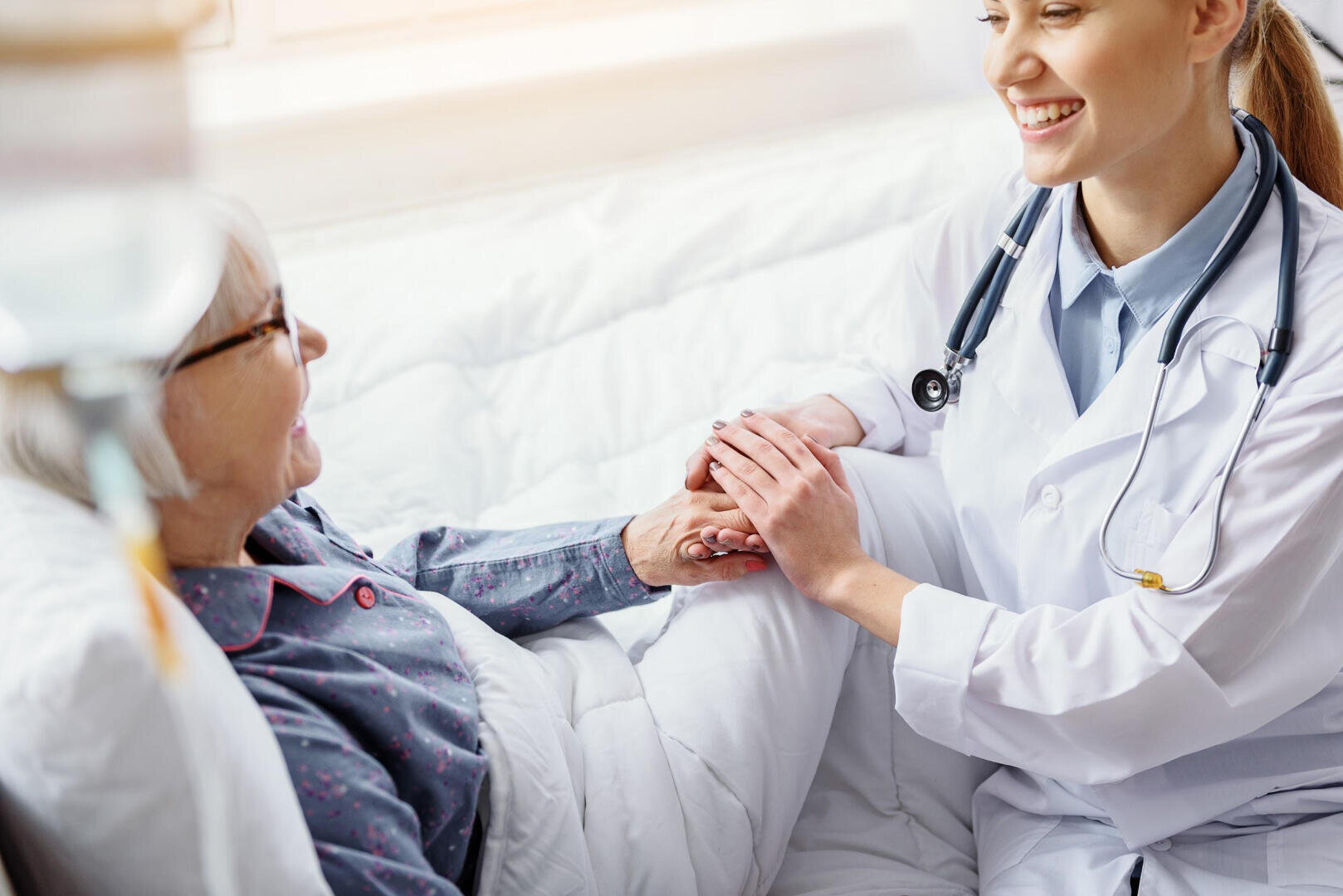 Ein lächelnder Arzt im weißen Kittel hält die Hand eines älteren Patienten, der in einem Krankenhausbett liegt.