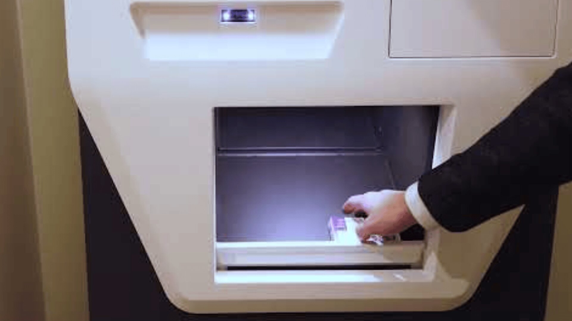 Eine Person in einem Anzug greift in das offene Fach eines Ein- oder Auszahlungsautomaten, möglicherweise in einer Bank oder einer gesicherten Einrichtung.