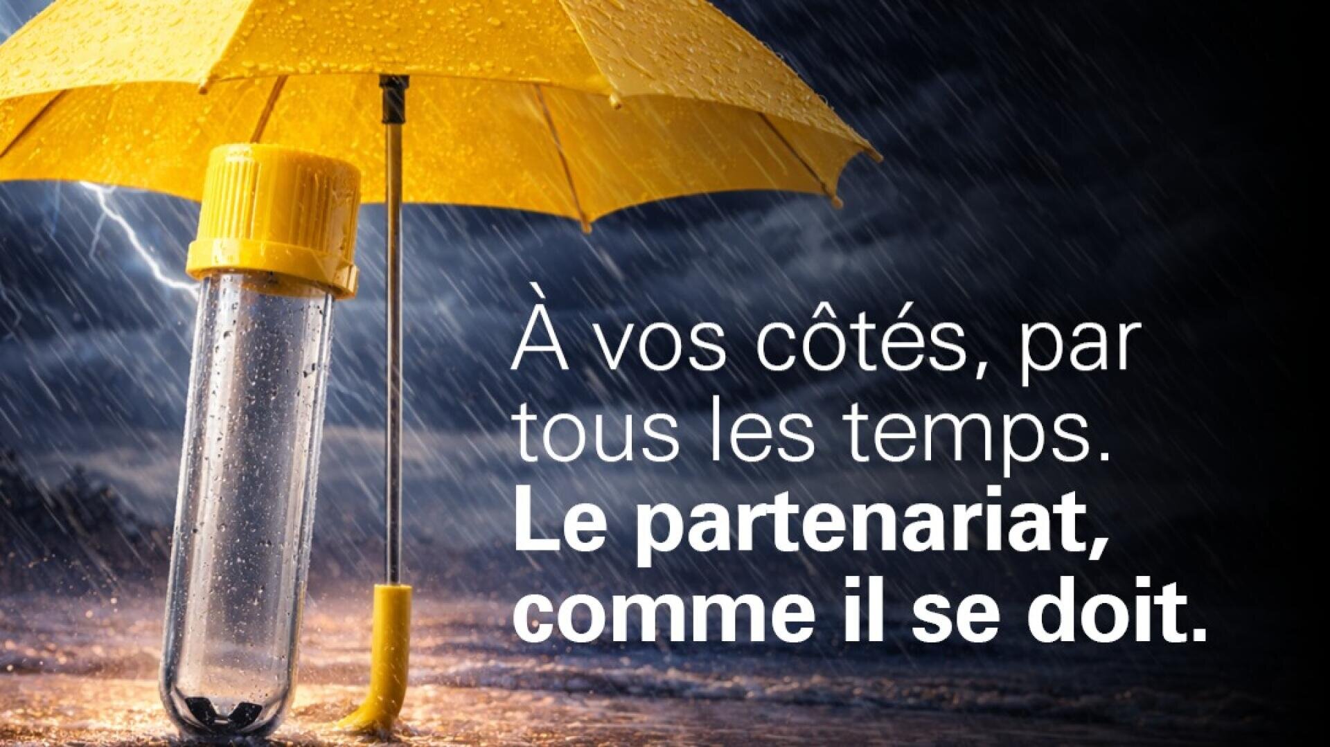Un parapluie jaune protège un tube à essai de laboratoire pendant un orage. On peut y lire en français : À vos côtés, par tous les temps. Le partenariat, comme il se doit.