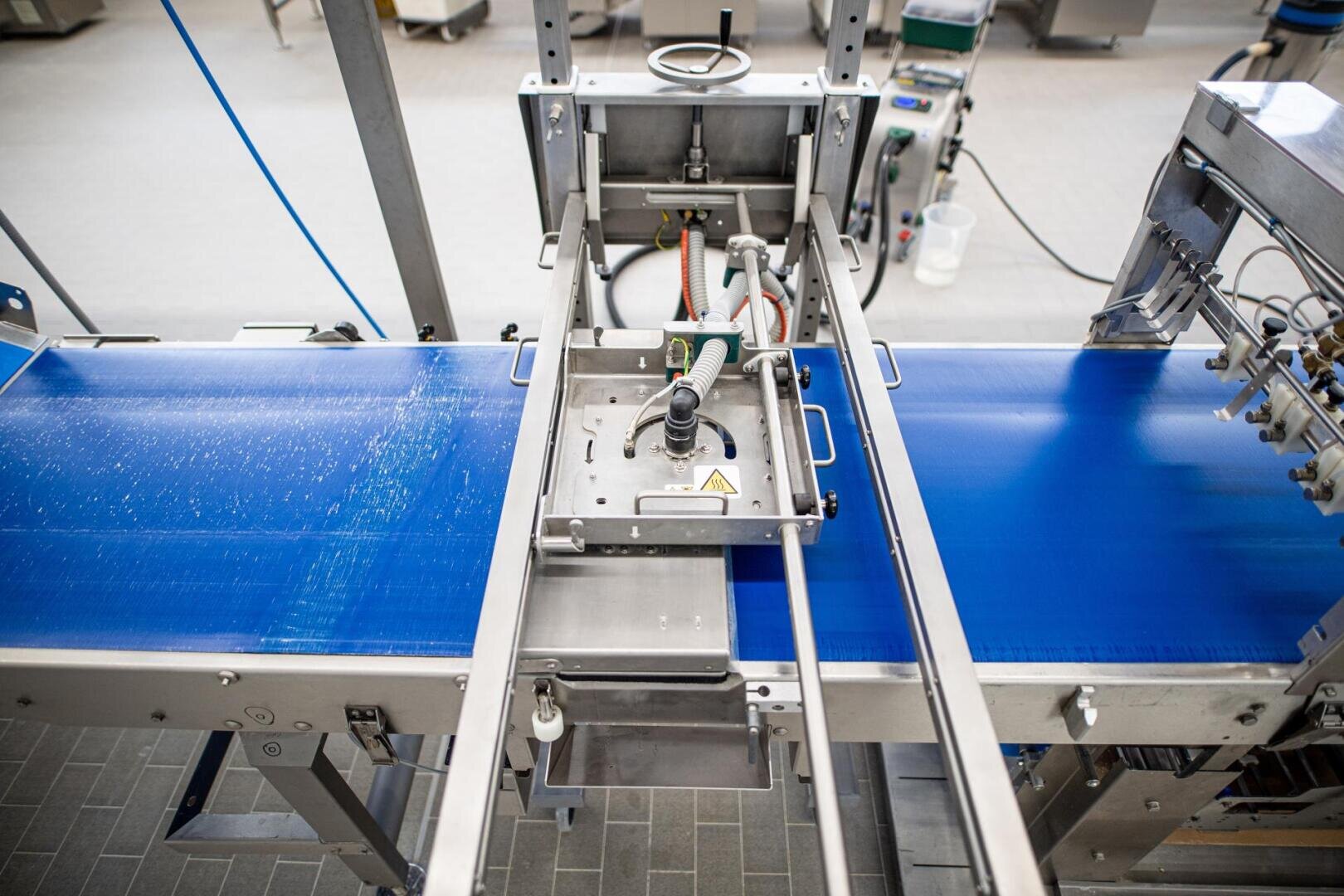 Draufsicht auf ein Förderbandsystem zur Lebensmittelverarbeitung in einer Fabrik mit Metallmaschinen, blauen Bändern, Schläuchen und einem Bedienfeld für die automatisierte Produktion.