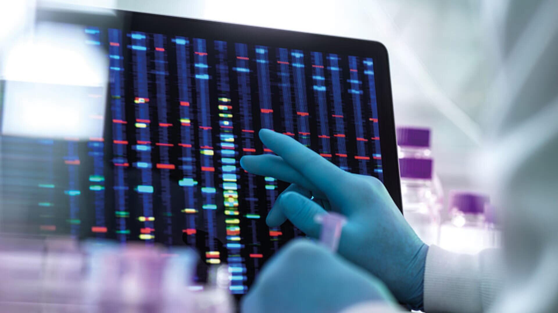 Eine Person mit blauen Handschuhen zeigt auf einen Computerbildschirm, auf dem eine bunte DNA-Sequenzierungsgrafik in einer Laborumgebung angezeigt wird, wobei Laborflaschen im Hintergrund verschwimmen.