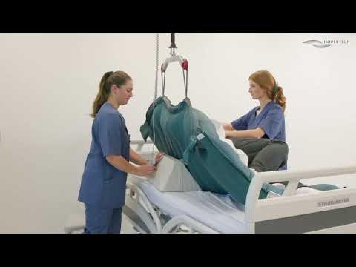 Zwei Mitarbeiter des Gesundheitswesens verwenden einen Deckenlifter mit einer Hebeschlinge, um einem Patienten dabei zu helfen, sich im Krankenhausbett aufzusetzen, und veranschaulichen so die Verfahren zur sicheren Patientenhandhabung im klinischen Umfeld.