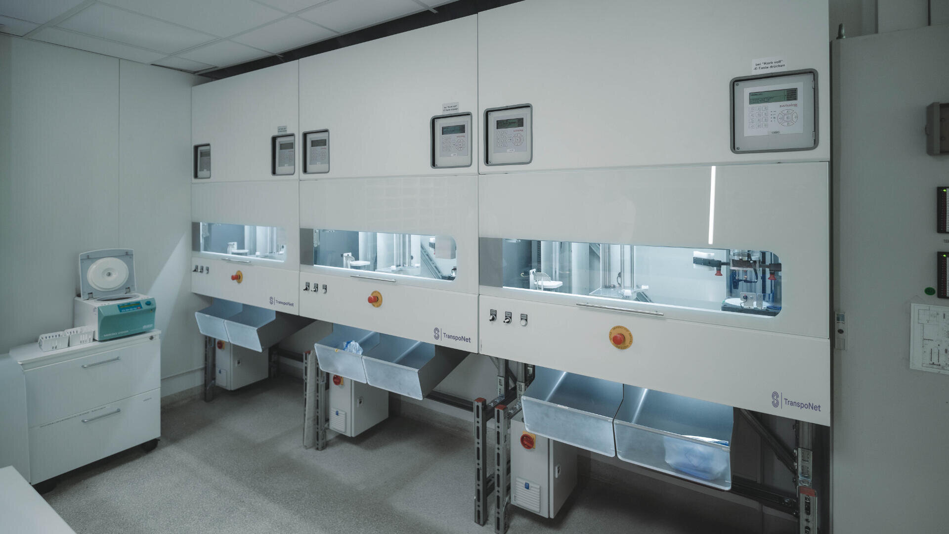 Ein modernes Labor mit vier automatisierten Arbeitsplätzen, digitalen Bedienfeldern und Ablagefächern unter jedem Arbeitsplatz. Der Raum ist sauber und übersichtlich, die Laborgeräte befinden sich auf der linken Seite.
