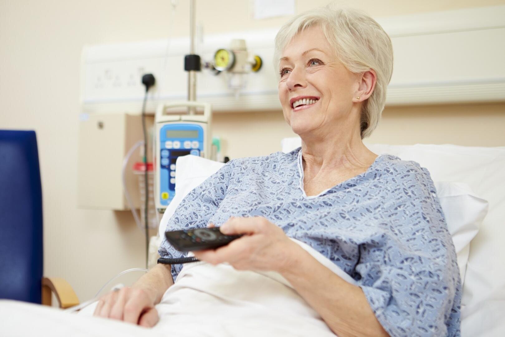 Eine ältere Frau in einem Krankenhausbett, die einen Kittel trägt, lächelt und hält eine Fernbedienung in der Hand. Neben ihr sind medizinische Geräte zu sehen, und sie wirkt zufrieden und entspannt.