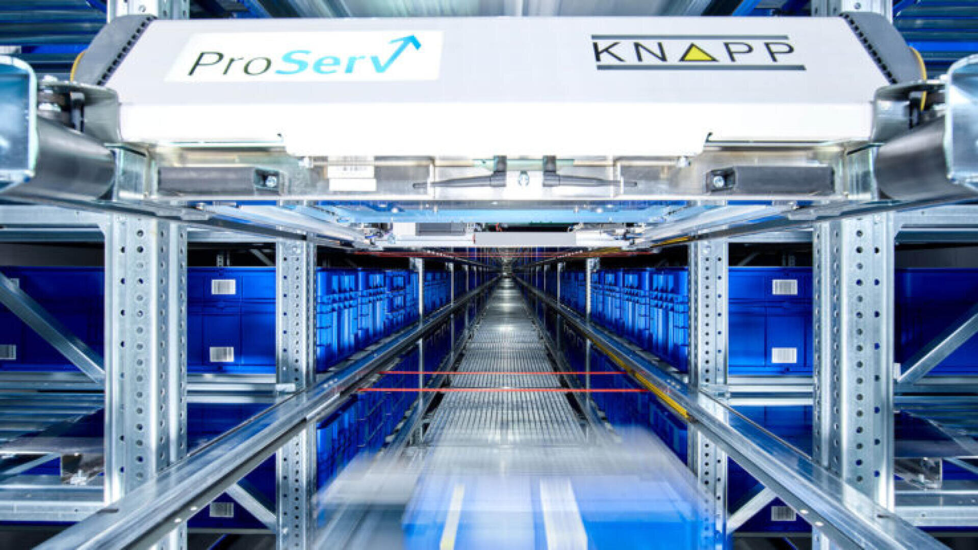 Ein automatisiertes Lagersystem mit Metallregalen, blauen Lagerbehältern und einem Roboter-Shuttle mit der Aufschrift „ProServ“ sowie einem „KNAPP“, das sich auf Schienen in einer hellen, modernen Halle fortbewegt.