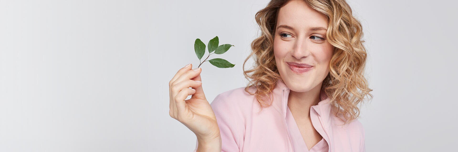Une femme vêtue d'un haut rose regarde une petite branche de feuilles qu'elle tient entre ses doigts.