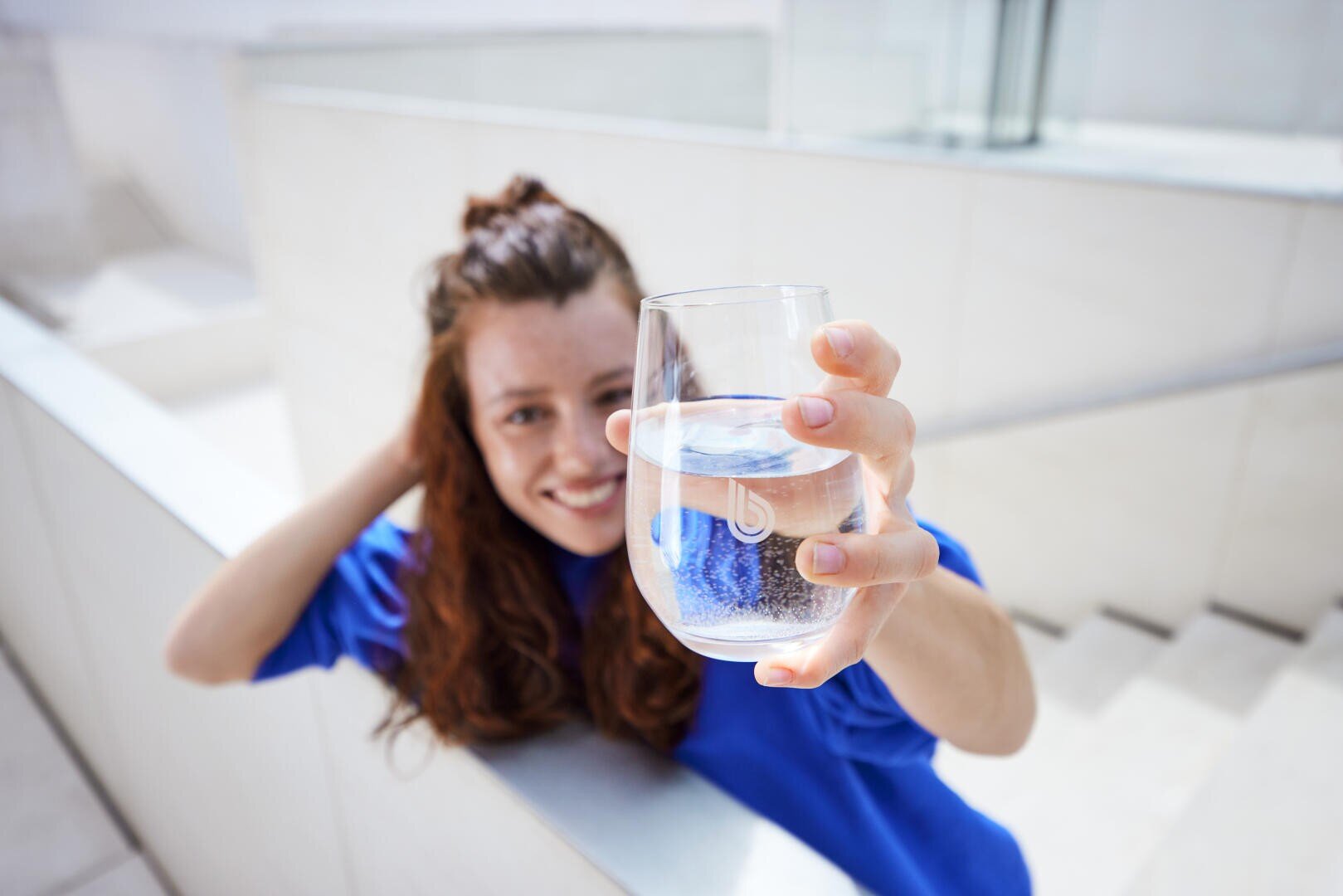 Eine Person mit langen, rötlichen Haaren, die ein blaues Hemd trägt, lächelt und hält ein Glas mit klarem Wasser in Richtung Kamera. Der Hintergrund ist hell und schlicht, mit weißen Wänden und einer Treppe.