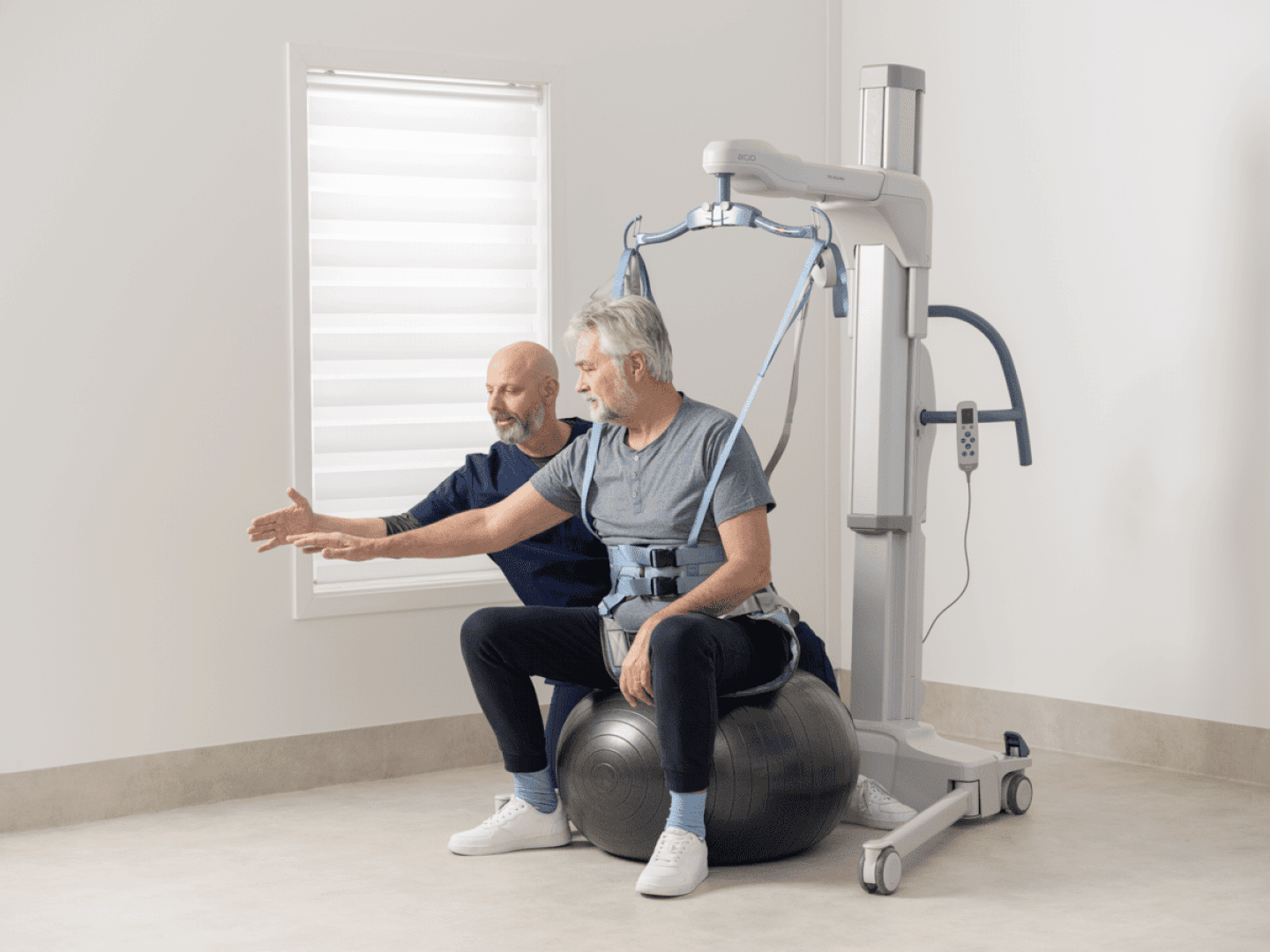 Ein älterer Mann sitzt auf einem Gymnastikball, an dem ein Gurt befestigt ist, der mit einem Patientenlifter verbunden ist, während ein Therapeut neben ihm kniet und seine Armbewegungen in einem hellen Behandlungsraum anleitet.
