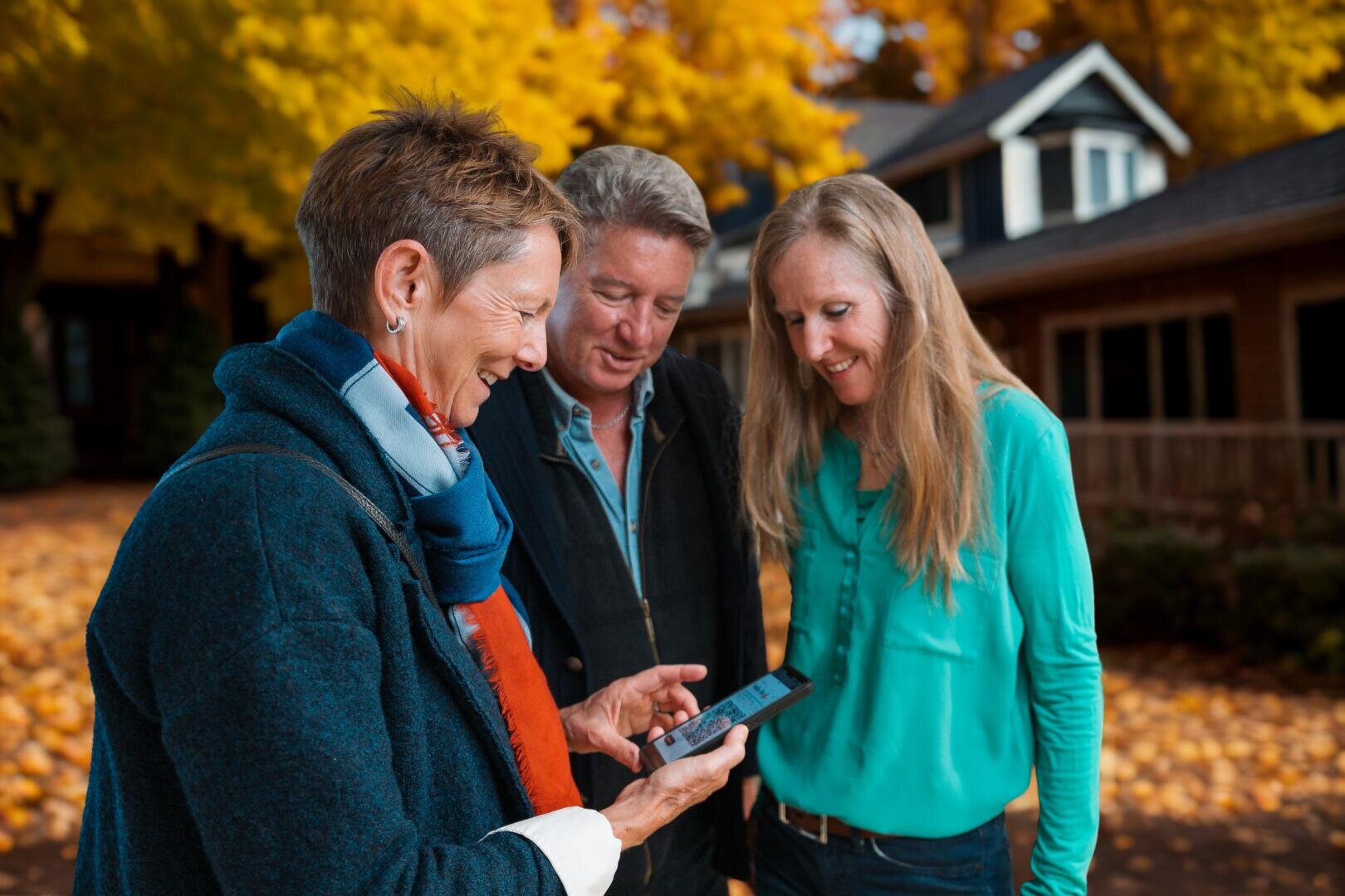 Trois adultes se tiennent dehors, sur un chemin recouvert de feuilles d'automne. Ils sourient et sont rassemblés autour d'un smartphone ; on dirait qu'ils regardent quelque chose ensemble. À l'arrière-plan, on aperçoit des arbres aux feuilles jaunes et une maison.