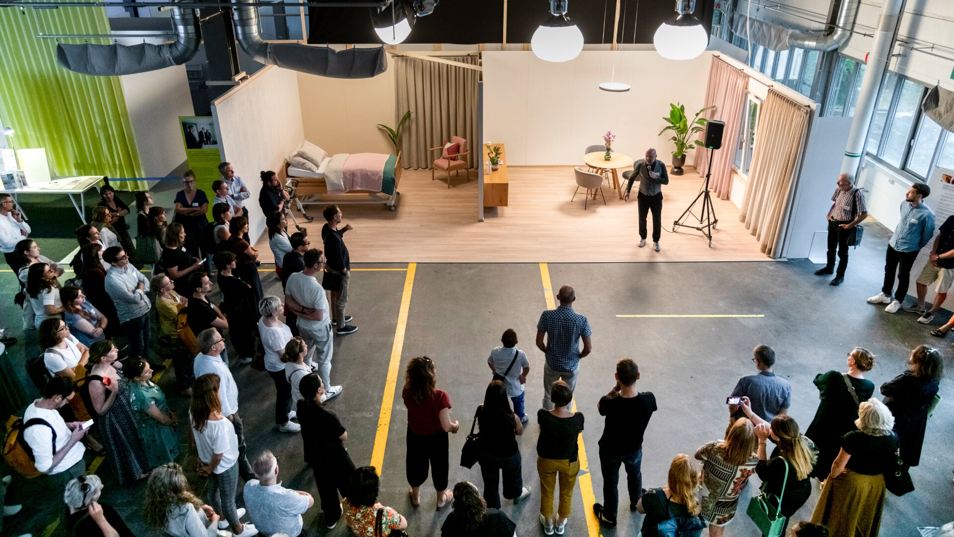 Eine große Gruppe von Menschen versammelt sich in einer Lagerhalle und schaut einem Redner zu, der mit einem Mikrofon vor einem inszenierten Wohnraum mit Bett, Schreibtisch und Esstisch steht, der hell erleuchtet ist.