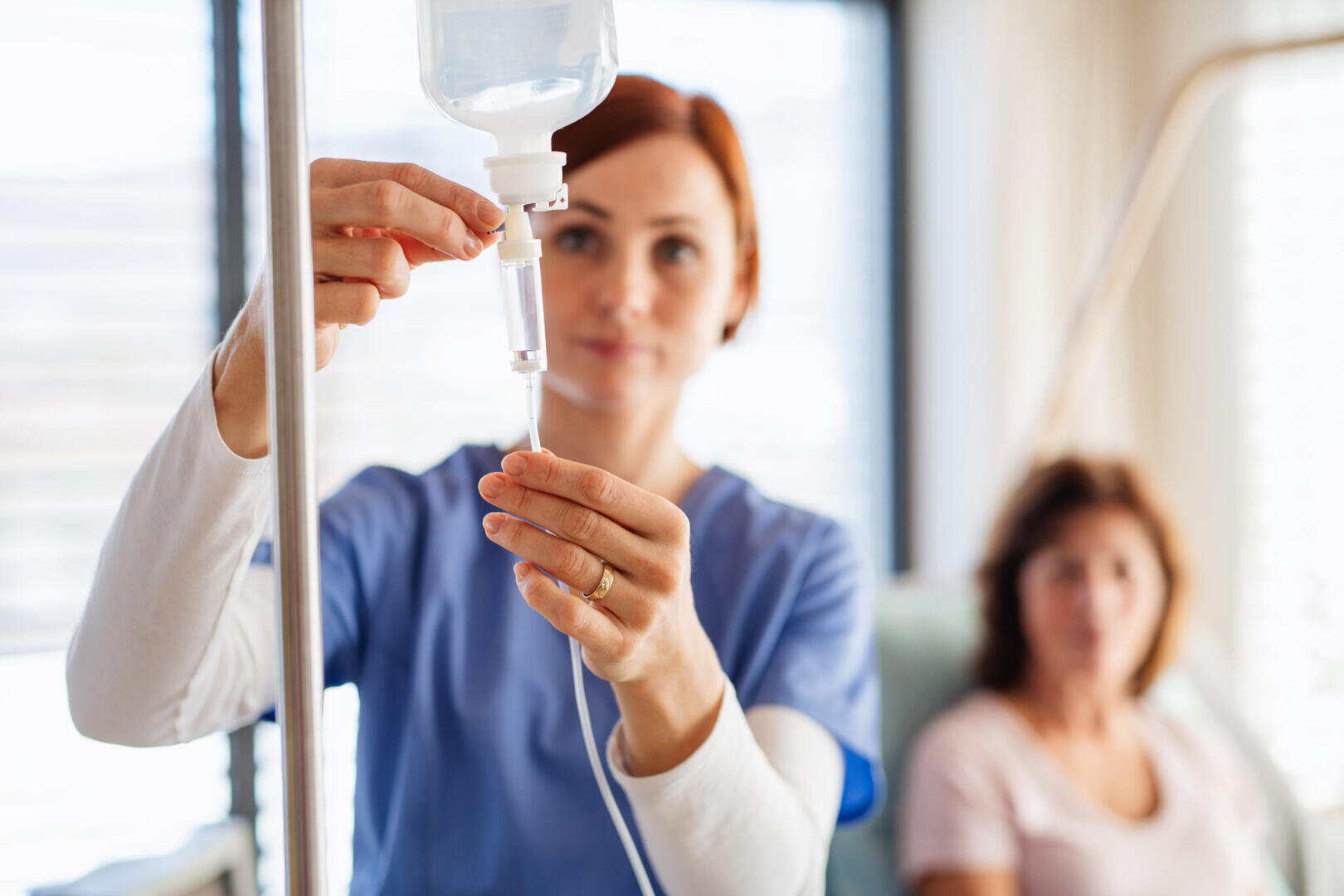 Ein medizinisches Fachpersonal befestigt einen Infusionsbeutel in einem Zimmer, in dem im Hintergrund ein Patient sitzt.