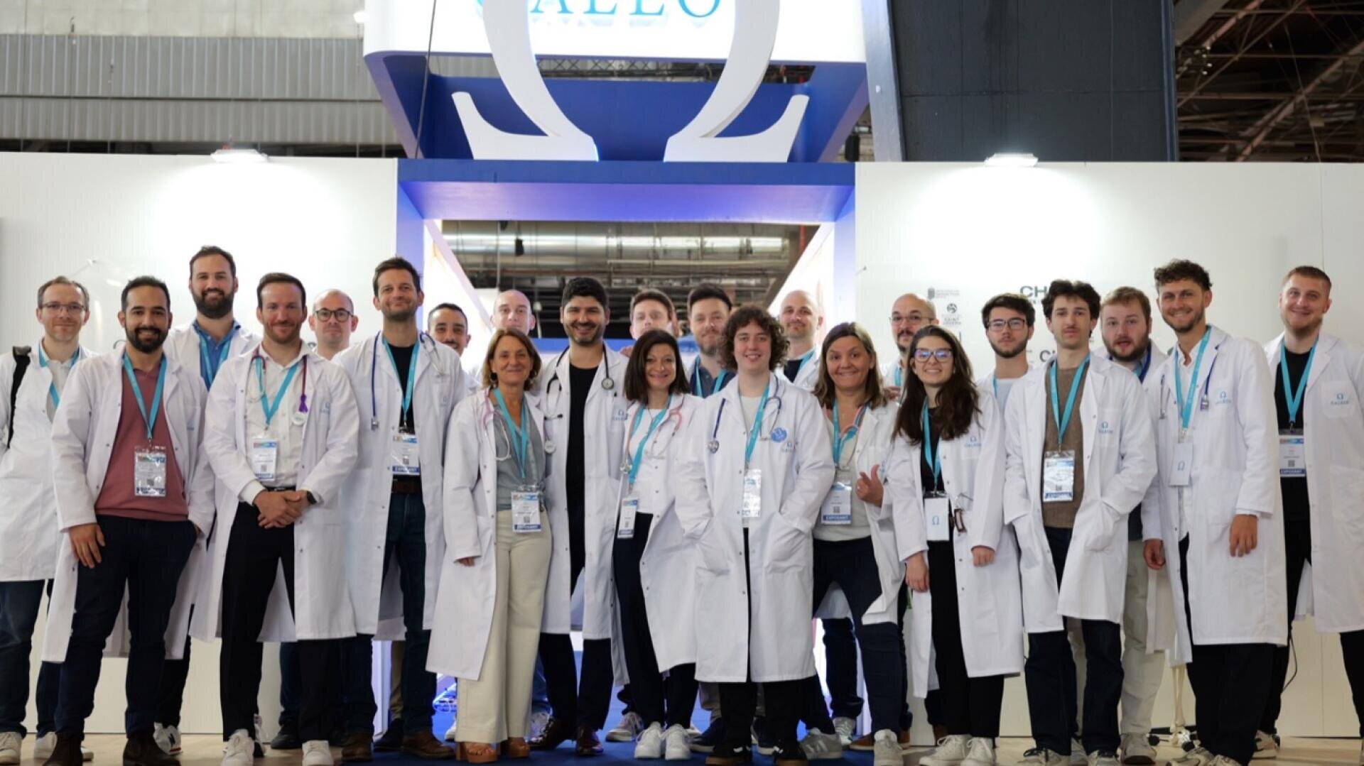 Un groupe de personnes vêtues de blouses blanches et portant des badges nominatifs posent ensemble en souriant pour une photo de groupe lors d'un événement ou d'une conférence en salle.