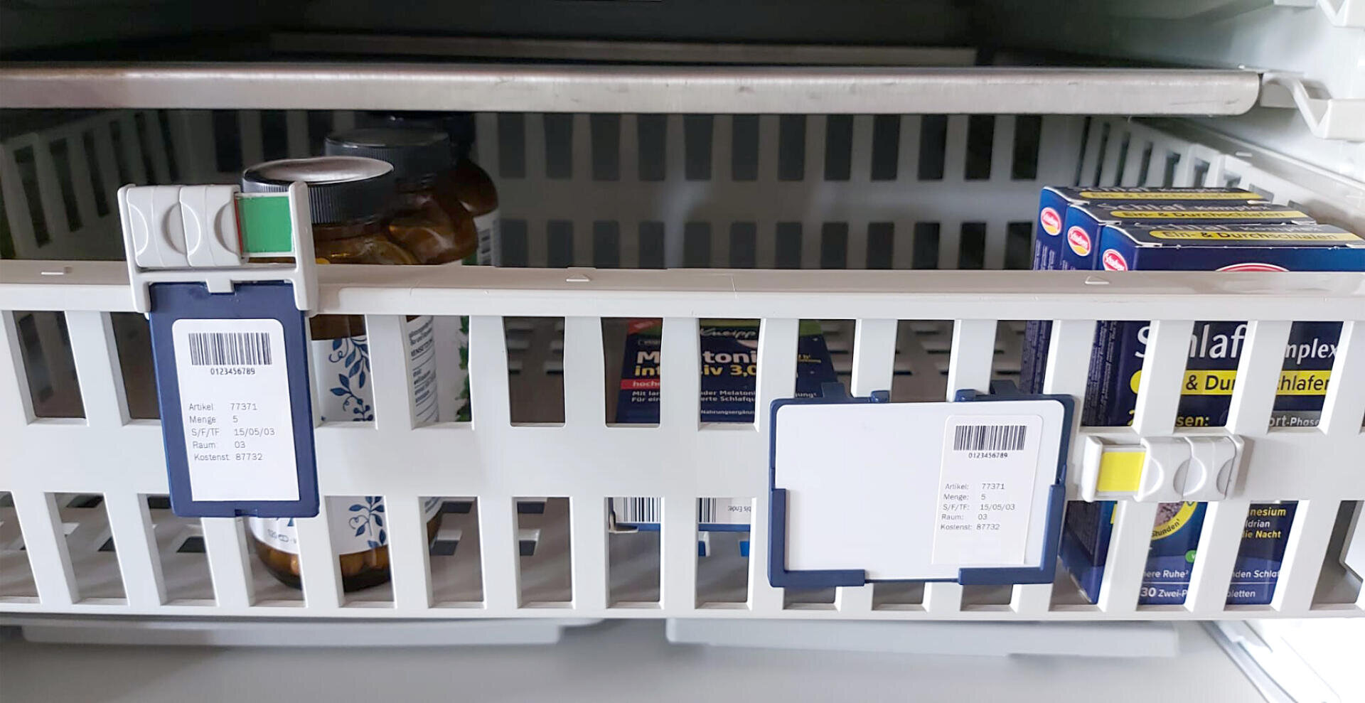 Dans un bac de rangement en plastique blanc doté de compartiments étiquetés se trouvent divers flacons et boîtes de médicaments, partiellement visibles derrière les lamelles ; le bac est placé dans un réfrigérateur ou une armoire de rangement.