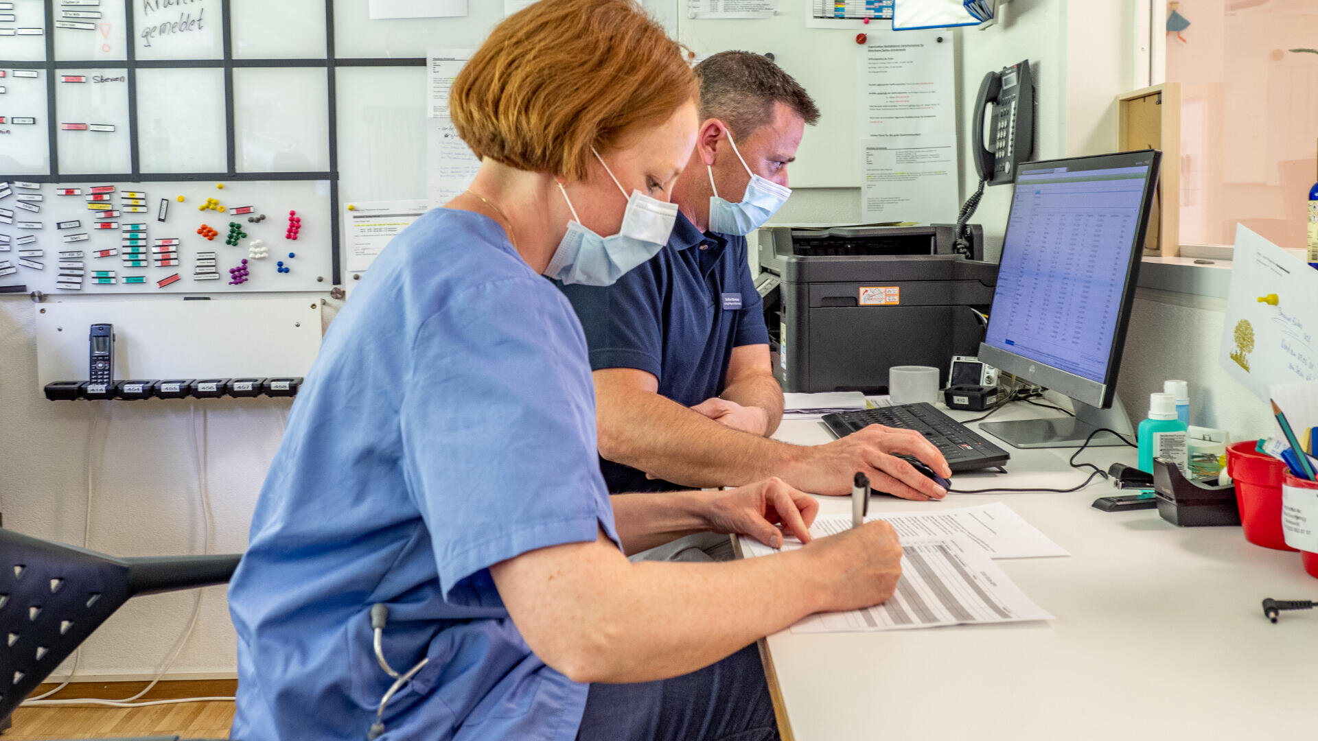 Zwei medizinische Fachkräfte, die Masken tragen, sitzen an einem Schreibtisch; einer schreibt an einem Papierkram, während der andere an einem Computer arbeitet, umgeben von Büromaterial und medizinischen Dokumenten.