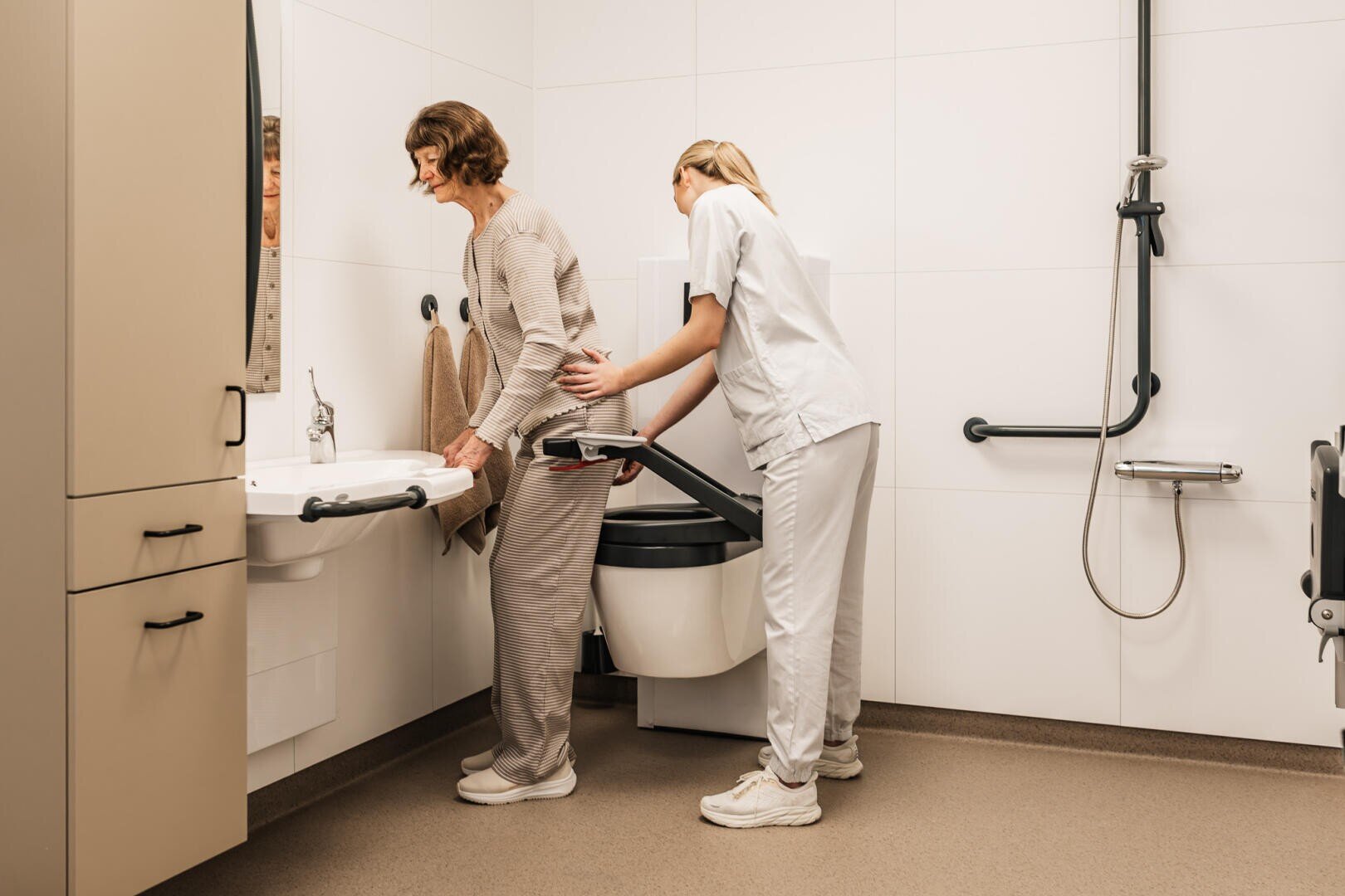 Ein Pflegekraft hilft einer älteren Frau vor einem Waschbecken in einem barrierefreien Badezimmer, das mit Haltegriffen und einer Dusche ausgestattet ist, wobei der Schwerpunkt auf Unterstützung und Mobilität liegt.