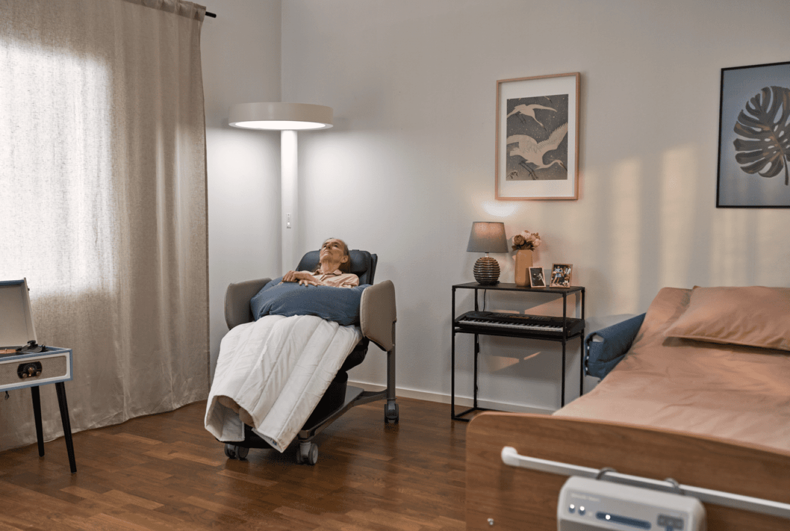 Ein älterer Mensch ruht sich in einem Liegesessel mit einer Decke in einem gemütlichen, sanft beleuchteten Schlafzimmer aus. Der Raum ist mit einem Bett, einem Beistelltisch, einer Lampe, Fotos und Wandbildern ausgestattet, was eine ruhige, behagliche Atmosphäre schafft.