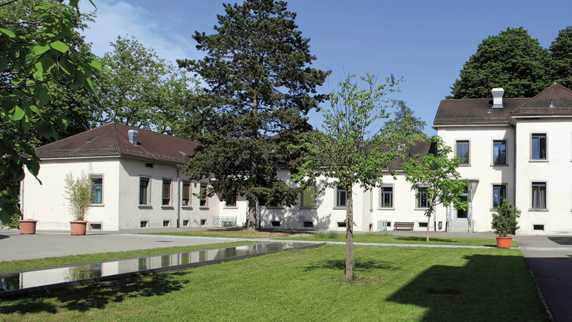 Ein weißes Gebäude mit einem braunen Dach steht inmitten von grünem Gras und Bäumen unter einem blauen Himmel, mit großen Fenstern und einem kleinen rechteckigen, spiegelnden Pool im Innenhof.