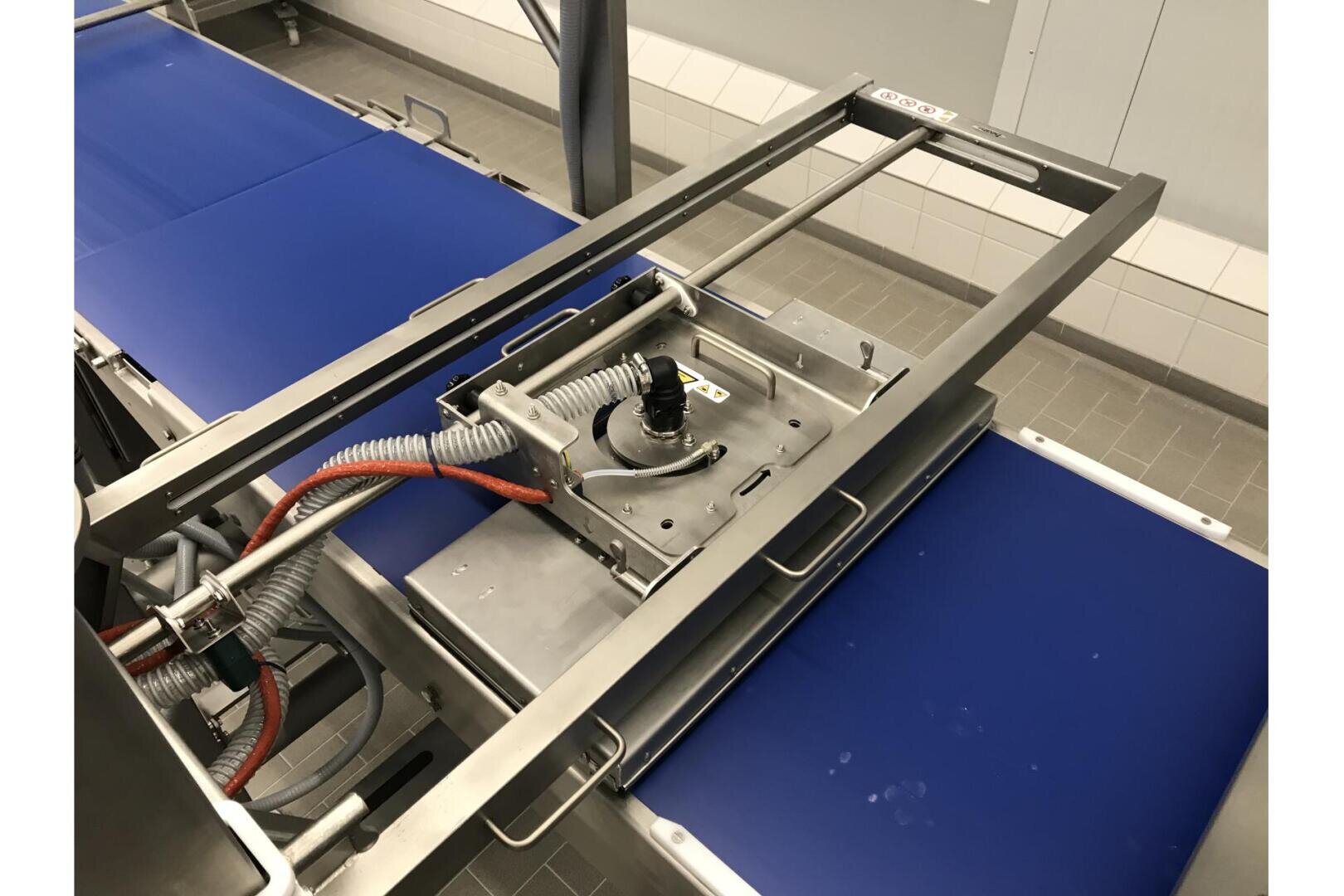 Eine Nahaufnahme von industriellen Förderbändern mit einem metallischen Maschinenteil in der Mitte, mit elektrischen Kabeln und einem schwarzen Knopf, in einer sauberen, gefliesten Produktionsumgebung.
