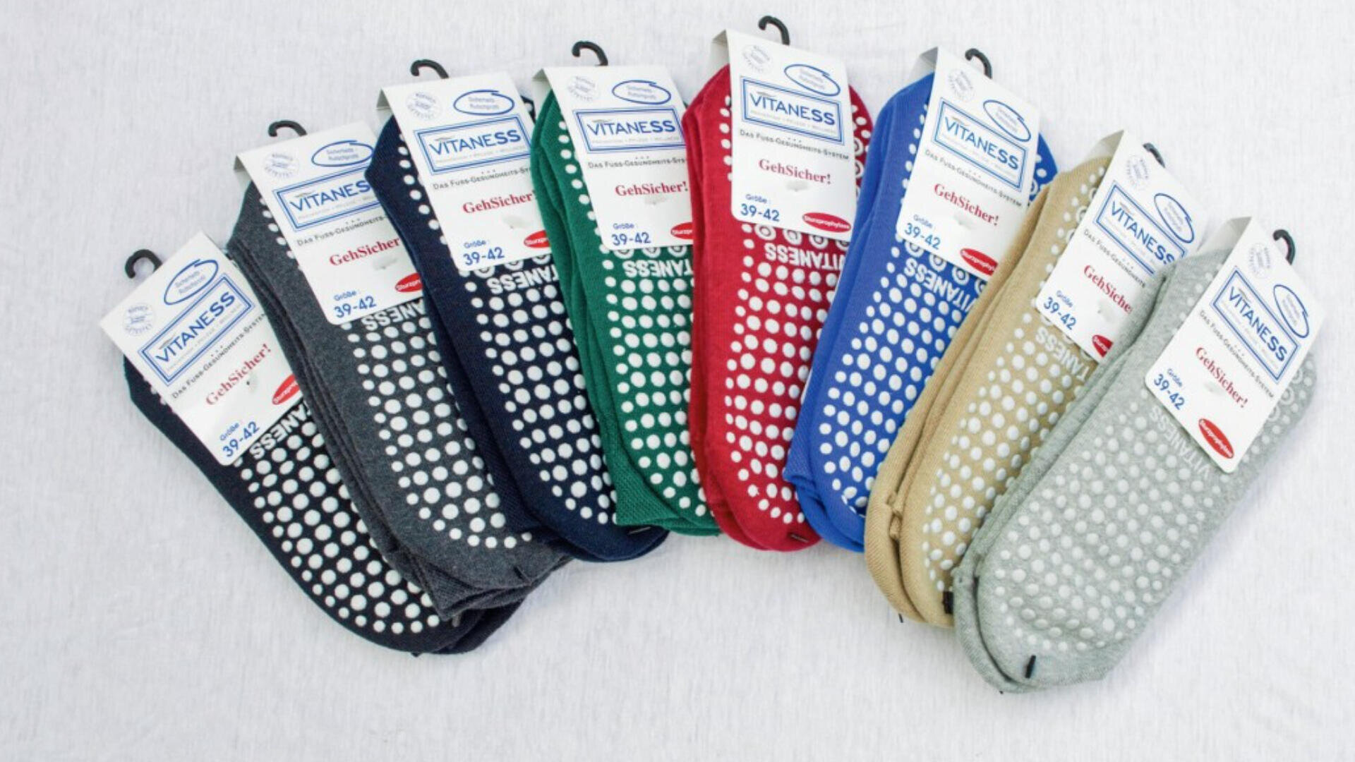 Acht Paar Vitaness-Antirutschsocken in verschiedenen Farben - schwarz, dunkelgrau, marineblau, grün, rot, blau, beige und hellgrau - sind in einer Reihe angeordnet und haben jeweils weiße Griffpunkte an den Sohlen.
