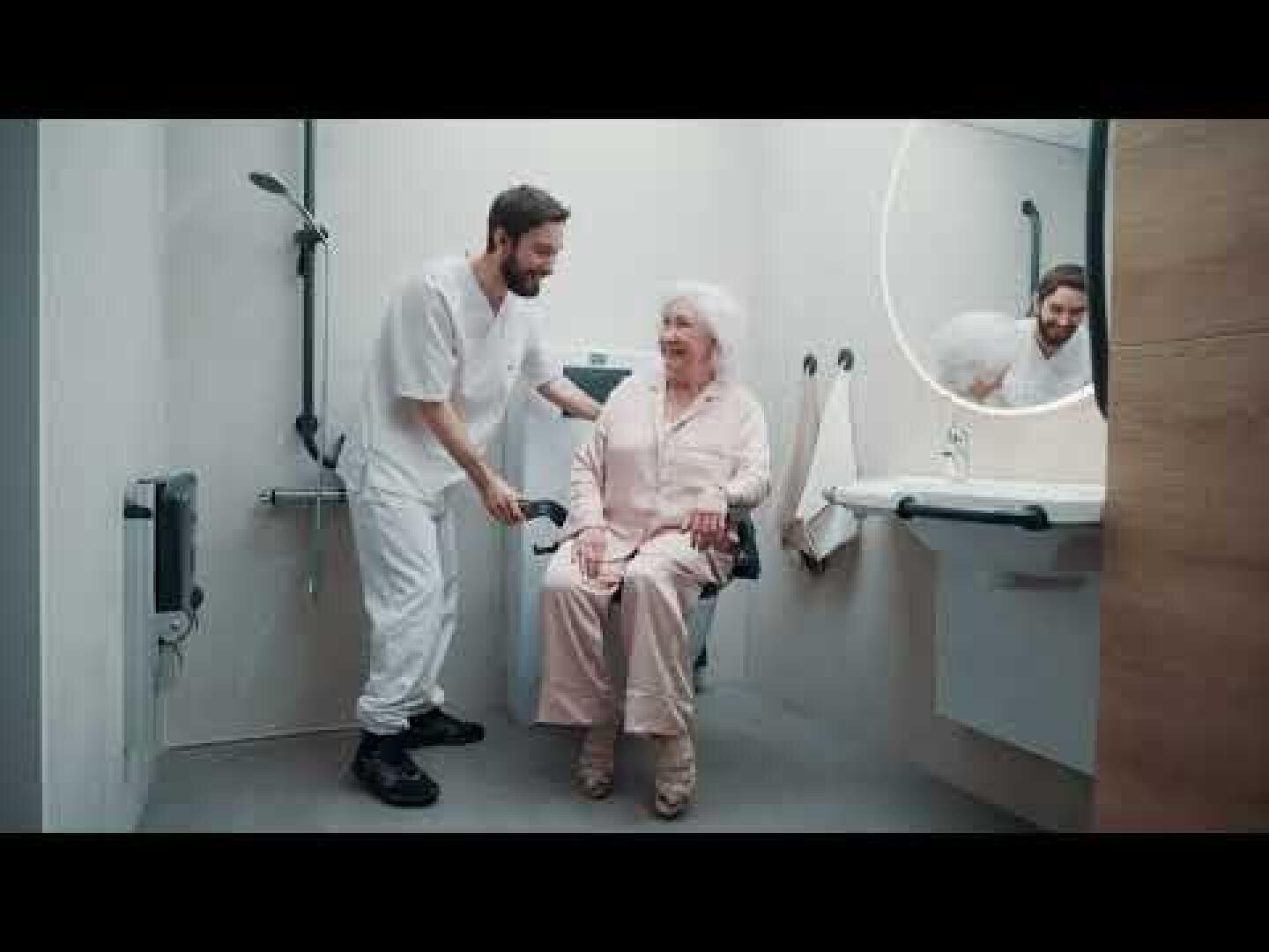 Un soignant aide une femme âgée assise sur une chaise de douche dans une salle de bains adaptée ; le reflet d'une autre personne apparaît dans le miroir près du lavabo.