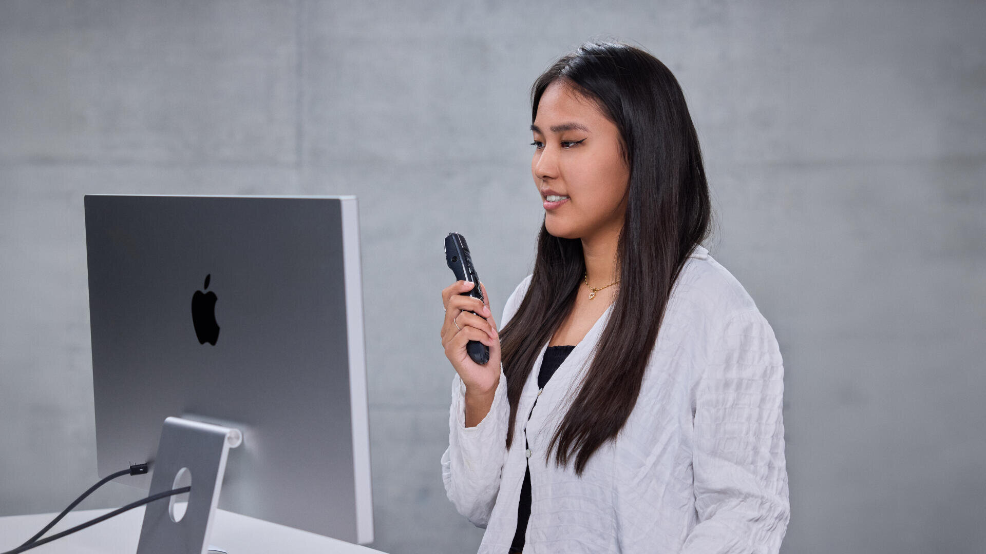 Une femme aux longs cheveux noirs tient un micro et parle devant un ordinateur de bureau Apple sur un fond gris uni.