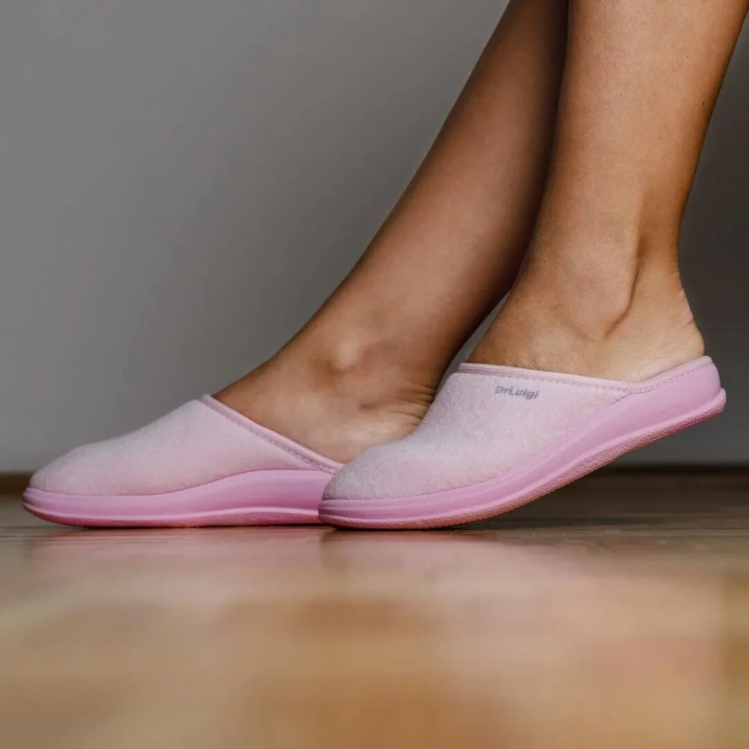 Eine Person trägt hellrosa Slipper und steht auf einem Holzboden vor einem schlichten grauen Hintergrund.