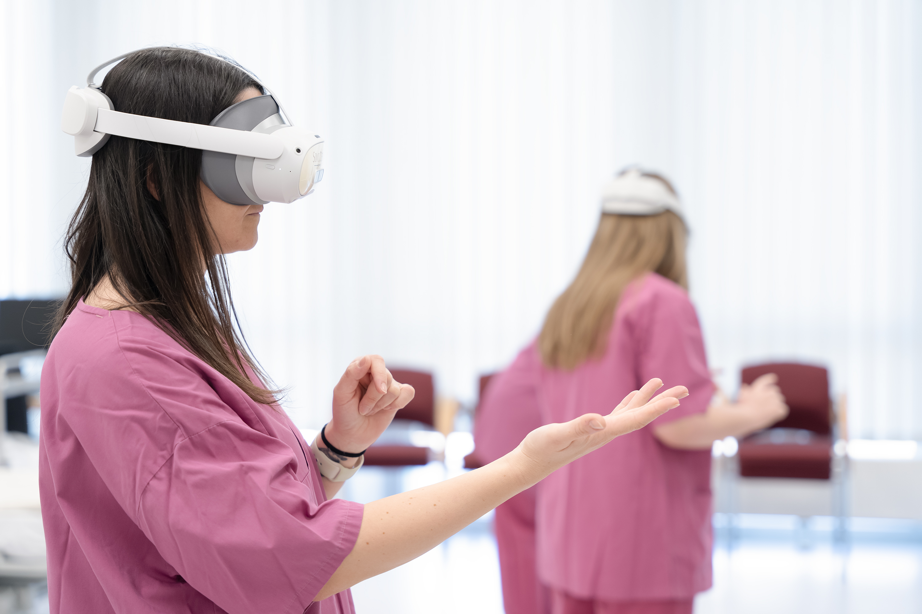Zwei Personen, die VR-Headsets tragen und rosa OP-Kleidung anhaben, gestikulieren mit den Händen.
