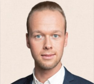 Eine Frau mit kurzen, hellbraunen Haaren lächelt in die Kamera. Sie trägt einen dunklen Blazer und kleine Ohrringe vor einem unscharfen, hellen Hintergrund.