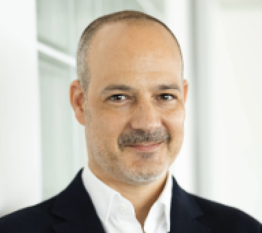 Eine Frau mit kurzen, hellbraunen Haaren lächelt in die Kamera. Sie trägt einen dunklen Blazer und kleine Ohrringe vor einem unscharfen, hellen Hintergrund.