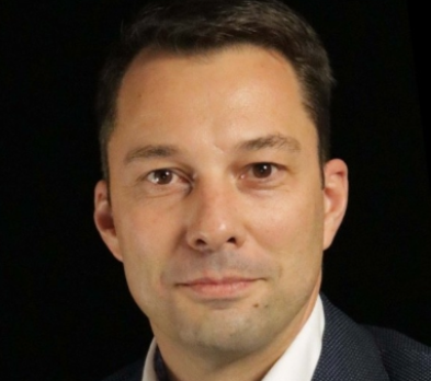 Ein Mann mit kurzen dunklen Haaren und leichtem Bartstoppeln posiert in einem dunklen Blazer vor einem hellen Holzhintergrund. Sein Gesichtsausdruck ist neutral und er blickt leicht zur Seite.