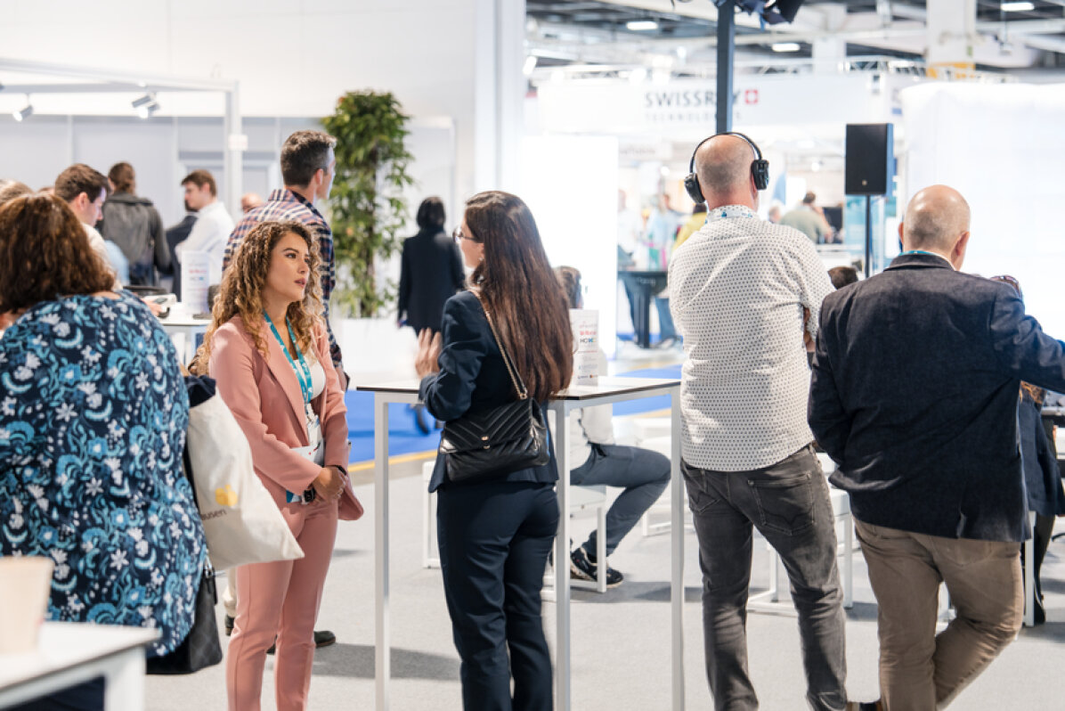 Un groupe de personnes se tient debout et discute lors d'un événement de réseautage ou d'une conférence dans un espace intérieur lumineux avec des stands et des écrans d'information en arrière-plan.